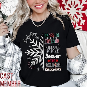 May include: Black t-shirt with a white snowflake design and text that reads "Kidsley Gray" and "Babes in Toyland 2024". The snowflake design includes the names of various Christmas characters, such as "Gingerbread Girl", "Jack in the Box", and "Soldier". The text "Prelude Icicle Jester Tap Soldier Chocolate" is also on the shirt.
