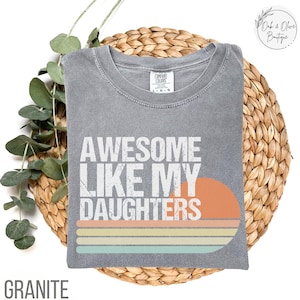 May include: A gray Comfort Colors t-shirt with the text "AWESOME LIKE MY DAUGHTERS" in white. The design includes an orange sun and horizontal stripes in orange, yellow, and blue. The word "GRANITE" is at the bottom.