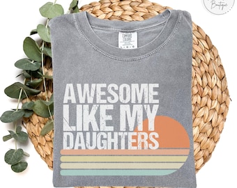 May include: A gray Comfort Colors t-shirt with the text "AWESOME LIKE MY DAUGHTERS" in white. The design includes an orange sun and horizontal stripes in orange, yellow, and blue. The word "GRANITE" is at the bottom.