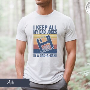 May include: A white t-shirt with a retro-style graphic of a floppy disk and the text "I keep all my dad jokes in a dad-a-base".