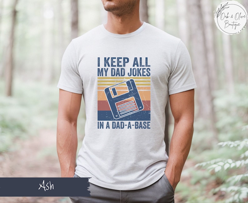 May include: A white t-shirt with a retro-style graphic of a floppy disk and the text "I keep all my dad jokes in a dad-a-base".