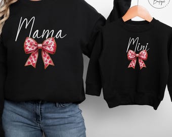 May include: Matching black sweatshirts with a white heart bow design. The larger sweatshirt says "Mama" and the smaller sweatshirt says "Mini".