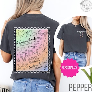 May include: A black t-shirt with a rainbow-colored graphic of school supplies and the text "Administration", "ABC", "Custom School Name", and a musical note. The graphic is in a postage stamp shape.