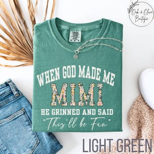 Puede incluir: Camiseta verde claro con el texto "WHEN GOD MADE ME MIMI HE GRINNED AND SAID 'This'll be Fun'". La palabra "MIMI" tiene un estampado floral. Un collar plateado est&aacute; sobre la camiseta. La camiseta est&aacute; doblada.