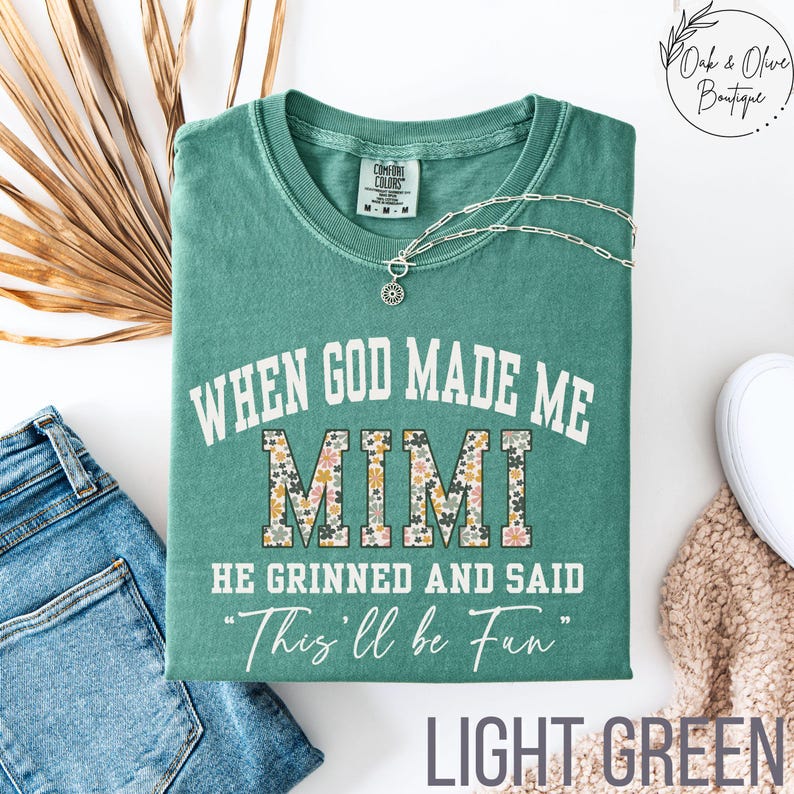 Puede incluir: Camiseta verde claro con el texto "WHEN GOD MADE ME MIMI HE GRINNED AND SAID 'This'll be Fun'". La palabra "MIMI" tiene un estampado floral. Un collar plateado est&aacute; sobre la camiseta. La camiseta est&aacute; doblada.