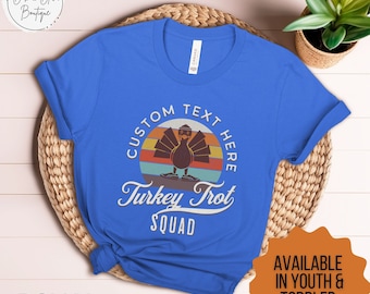 May include: A royal blue t-shirt with a retro-style design featuring a turkey wearing a hat and running. The text "CUSTOM TEXT HERE" is above the turkey, and the text "Turkey Trot SQUAD" is below the turkey. The text "AVAILABLE IN YOUTH & TODDLER SIZES TOO" is in the lower right corner of the image.