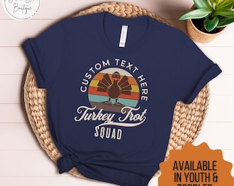 May include: Navy blue t-shirt with a vintage-style graphic of a turkey in front of a sunset. The text "CUSTOM TEXT HERE" is above the turkey and the text "Turkey Trot SQUAD" is below the turkey.