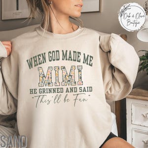 Puede incluir: Sudadera de color arena con el texto "WHEN GOD MADE ME MIMI HE GRINNED AND SAID 'This'll be Fun'" en verde y estampado floral. La sudadera tiene mangas largas y cuello redondo. El texto "Oak & Olive Boutique" est&aacute; en un c&iacute;rculo.