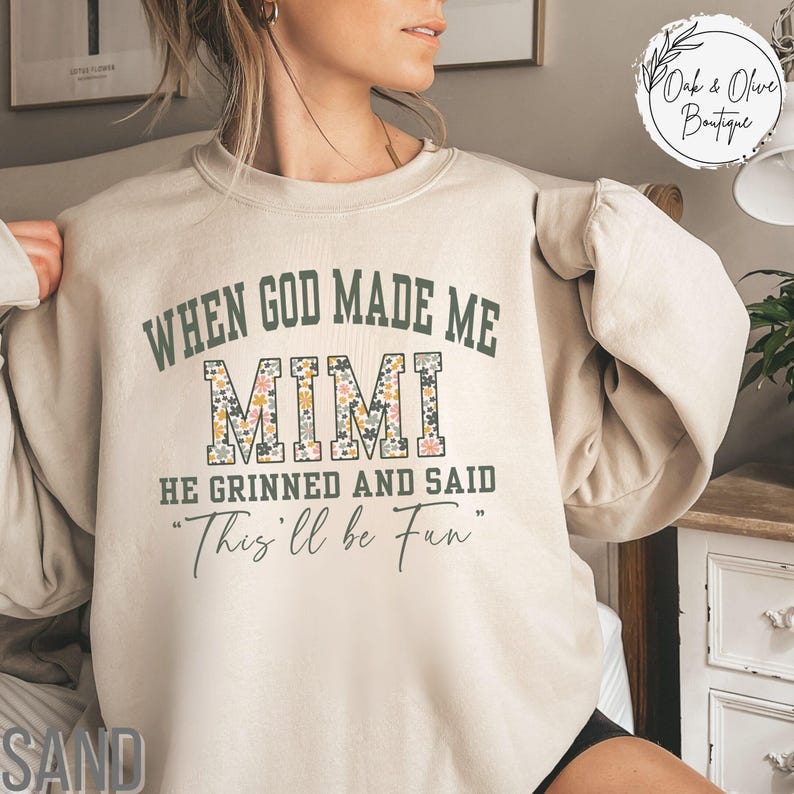 Puede incluir: Sudadera de color arena con el texto "WHEN GOD MADE ME MIMI HE GRINNED AND SAID 'This'll be Fun'" en verde y estampado floral. La sudadera tiene mangas largas y cuello redondo. El texto "Oak & Olive Boutique" est&aacute; en un c&iacute;rculo.