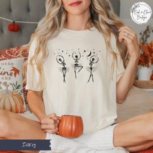 May include: A white t-shirt with a graphic of three skeletons dressed as ballerinas dancing under a crescent moon and stars. The text "Oak & Olive Boutique" is in the upper right corner of the image. The t-shirt is on a model wearing it. The model is sitting on a couch with a pumpkin mug in her hand.