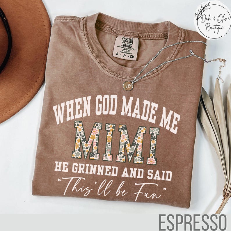 Puede incluir: Camiseta color espresso con el texto "WHEN GOD MADE ME MIMI HE GRINNED AND SAID 'This'll be Fun'" en blanco y estampado floral. La camiseta tiene cuello redondo y mangas cortas. Un collar plateado con un colgante est&aacute; drapeado sobre la camiseta.