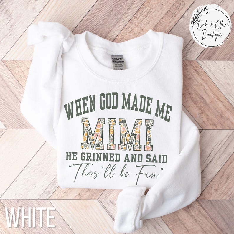Puede incluir: Sudadera blanca con el texto "WHEN GOD MADE ME MIMI HE GRINNED AND SAID 'This'll be Fun'" en verde y estampado floral. La palabra "WHITE" est&aacute; en la parte inferior. La sudadera est&aacute; doblada sobre una superficie de madera.