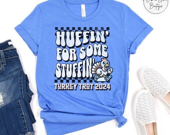 May include: A blue t-shirt with a white checkered background and the text "HUFFIN' FOR SOME STUFFIN' TURKEY TROT 2024". A cartoon turkey is running with a red and white hat and a red and white scarf.