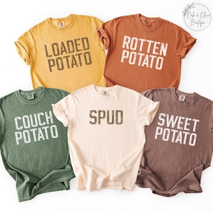 May include: Five colorful t-shirts with potato-themed text. The shirts are in shades of yellow, orange, green, cream, and brown. Text includes "LOADED POTATO", "ROTTEN POTATO", "COUCH POTATO", "SPUD", and "SWEET POTATO".