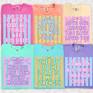 May include: A collection of six colorful t-shirts, each featuring a different inspirational quote in a decorative design. The shirts are in various pastel colors, with text in a contrasting color. The quotes include phrases like "With God all things are possible" and "Fear not for I am with you!"