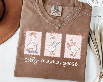 May include: Espresso-colored Comfort Colors t-shirt featuring three cartoon geese in pink frames. The geese are engaged in different activities, including riding a scooter, reading a book, and drinking coffee. The text "silly mama goose" is below the illustrations.