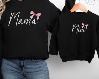 May include: Matching black sweatshirts with pink bows. The adult sweatshirt says "Mama" and the child's sweatshirt says "Mini".
