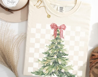 May include: Ivory-colored t-shirt with a Christmas tree design. The tree is decorated with white lights and topped with a red bow. The shirt has a checkered background and the text "IVORY" at the bottom.
