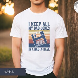 May include: White t-shirt with a retro-style graphic of a floppy disk and the text "I keep all my dad jokes in a dad-a-base".