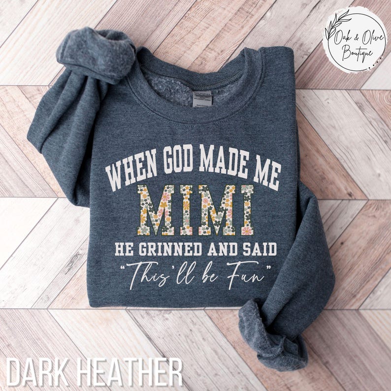Puede incluir: Sudadera de color heather oscuro con el texto "WHEN GOD MADE ME MIMI HE GRINNED AND SAID 'This'll be Fun'". La palabra "MIMI" tiene un estampado floral. La sudadera est&aacute; doblada sobre un fondo de madera clara.