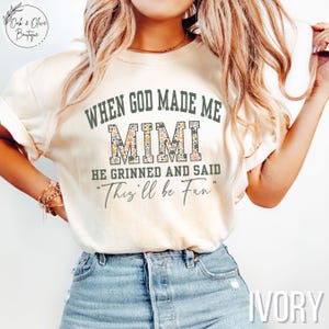 Puede incluir: Camiseta de color marfil con el texto "WHEN GOD MADE ME MIMI HE GRINNED AND SAID 'This'll be Fun'". La palabra "MIMI" est&aacute; rellena con un estampado floral. La camiseta se combina con vaqueros azules.