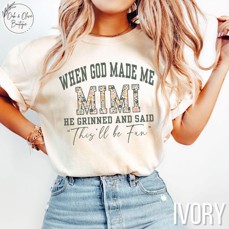 Puede incluir: Camiseta de color marfil con el texto "WHEN GOD MADE ME MIMI HE GRINNED AND SAID 'This'll be Fun'". La palabra "MIMI" est&aacute; rellena con un estampado floral. La camiseta se combina con vaqueros azules.