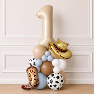 May include: A balloon arrangement featuring a large beige number one balloon, a gold cowboy hat balloon, and a brown cowboy boot balloon. The display includes round balloons in shades of beige, white, light blue, and white with black cow print patterns.