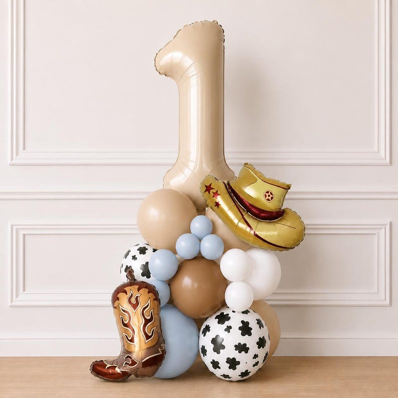 May include: A balloon arrangement featuring a large beige number one balloon, a gold cowboy hat balloon, and a brown cowboy boot balloon. The display includes round balloons in shades of beige, white, light blue, and white with black cow print patterns.