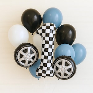 May include: A collection of balloons for a first birthday celebration. The arrangement includes black, white, and blue balloons, along with two tire-shaped balloons and a large number one balloon with a black and white checkered pattern.