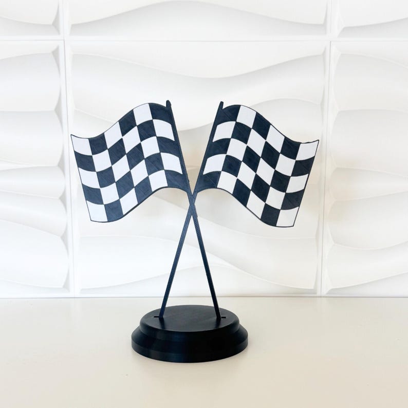 May include: A decorative tabletop display featuring two crossed checkered racing flags in black and white. The flags are mounted on thin black poles that are secured into a solid, circular black base, creating a stable structure for display.