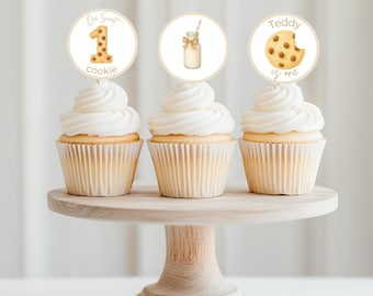 May include: Three cupcakes with white frosting and decorative toppers on a wooden cake stand. The toppers feature a number one, a milk bottle, and a cookie with text that reads "One Sweet Cookie" and "Teddy is one".