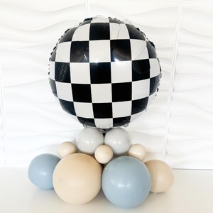 May include: A balloon arrangement with a large, round black and white chequered balloon at the top. The base has round balloons in white, light blue, and beige. The arrangement is set against a white background.