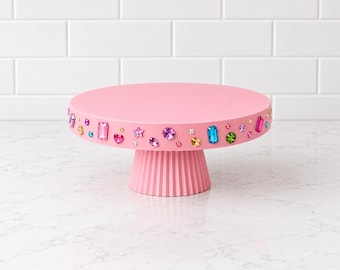 May include: A round, light pink cake stand with a fluted pedestal base. The vertical edge of the flat top is decorated with an assortment of colorful, multi-shaped plastic gemstones, including stars, hearts, and rectangles in shades of blue, green, and pink.