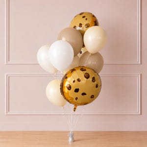 May include: A balloon bouquet featuring a mix of round latex balloons in shades of white, cream, and tan, alongside two round foil balloons printed with a brown chocolate chip cookie pattern, all tied together with white ribbons to a silver weight.