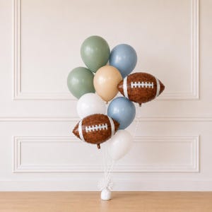 May include: A balloon bouquet featuring two brown football-shaped foil balloons with white laces, alongside round latex balloons in sage green, dusty blue, beige, and white, all tied together with thin white ribbons to a small white base.