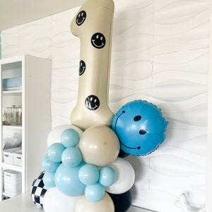 Pu&ograve; includere: Una composizione di palloncini che include un grande palloncino beige a forma di numero uno decorato con faccine sorridenti nere, un palloncino rotondo blu con faccina sorridente e un gruppo di palloncini pi&ugrave; piccoli bianchi, azzurri, beige e a scacchi.