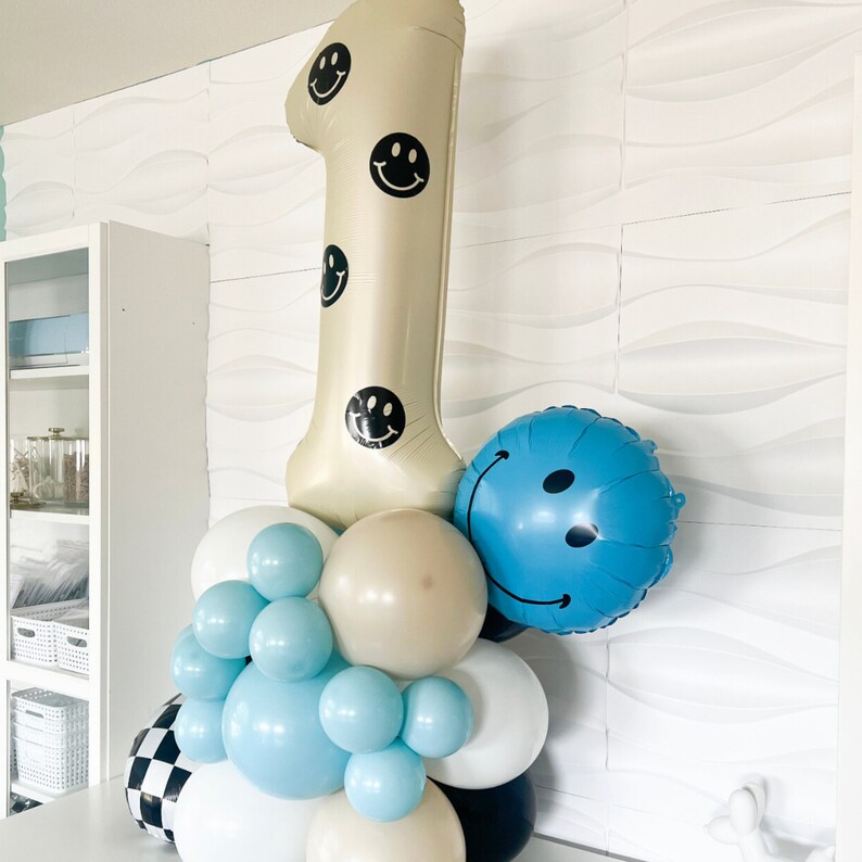 Pu&ograve; includere: Una composizione di palloncini che include un grande palloncino beige a forma di numero uno decorato con faccine sorridenti nere, un palloncino rotondo blu con faccina sorridente e un gruppo di palloncini pi&ugrave; piccoli bianchi, azzurri, beige e a scacchi.