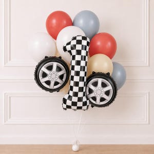 Puede incluir: Un ramo de globos que incluye un globo grande con forma de número uno a cuadros blancos y negros, dos globos con forma de neumático en negro y plata, y varios globos redondos en tonos rojo, blanco, gris y crema, unidos con cintas blancas.