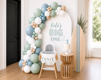May include: A first birthday party setup with a high chair, balloons, and a surfboard. The high chair has a banner that says "ONE". A cake with a number 1 on top sits on a small table. The backdrop has the text "Kole's BIG ONE".