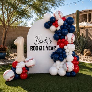 May include: A white arched backdrop with the black text Brody's ROOKIE YEAR, decorated with a balloon garland in navy blue, red, and white, including baseball-patterned balloons and a large beige number one balloon on the left side.
