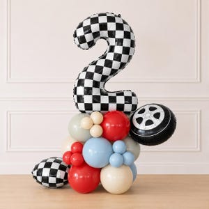 May include: A large foil balloon shaped like the number two with a black and white checkered pattern, supported by a cluster of round latex balloons in red, light blue, cream, and grey, plus a black and white checkered sphere and a tire balloon.