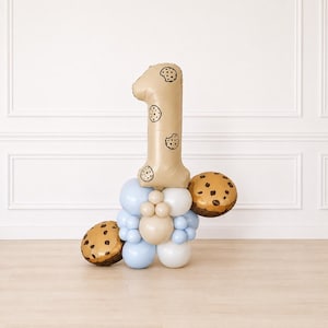 May include: A large beige number one balloon decorated with small black cookie illustrations, surrounded by a cluster of light blue, white, and beige round balloons, with two additional round cookie-patterned balloons on the sides.