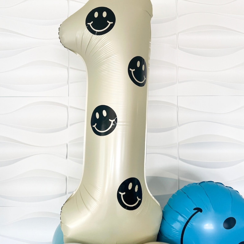 Pu&ograve; includere: Un grande palloncino in alluminio color beige a forma di numero uno, decorato con diverse icone di faccine sorridenti nere. Il palloncino presenta una finitura metallica liscia ed &egrave; posizionato accanto a una parte di un palloncino blu rotondo.