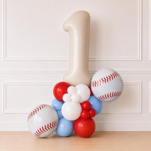 May include: A balloon arrangement featuring a large beige number one balloon as the centerpiece, flanked by two round white balloons with red baseball stitching patterns, accented by smaller red, white, and light blue round balloons.