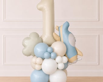 May include: A balloon arrangement featuring a large beige number one foil balloon, a blue and beige airplane foil balloon, and a cloud-shaped balloon. The base consists of a cluster of round latex balloons in shades of matte blue, white, and beige.