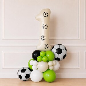 以下が含まれることがあります： 黒と白のサッカーボールのグラフィックが描かれたクリーム色の大きな数字の「1」のバルーン。ライムグリーン、黒、白、クリーム色の丸いバルーンの束の上に配置されており、その中には2つの球形のサッカーボールバルーンが含まれています。