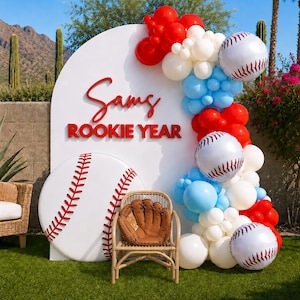 Puede incluir: Un panel arqueado blanco con el texto rojo 'Sams ROOKIE YEAR' y un gráfico de béisbol. Una silla de ratán con un guante de béisbol marrón se sitúa delante, decorada con una guirnalda de globos en rojo, blanco y azul claro.
