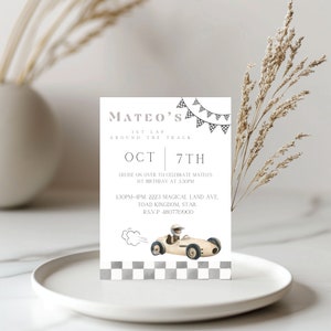 Puede incluir: Una invitación gris y blanca con un borde a cuadros. La invitación es para el primer cumpleaños de Mateo. El texto dice "Mateo's 1st Lap Around the Track Oct 7th. Cruise on over to celebrate Mateo's 1st birthday at 3:30pm. 1:30pm-4pm. 2223 Magical Land Ave. Toad Kingdom, Star. RSVP 4807719900". Una ilustración de un coche de carreras de juguete se encuentra en la esquina inferior derecha.