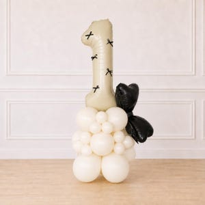 May include: A beige number one balloon featuring small black bow graphics, positioned atop a cluster of white balloons. A large black balloon shaped like a bow is attached to the side of the white balloon base, creating a decorative display.