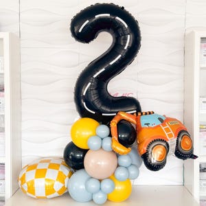 Puede incluir: Un globo negro con el número dos con líneas blancas, un globo de camión de construcción y un grupo de globos amarillos, azules y marrones.